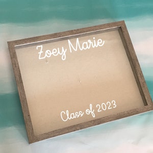 Puede incluir: Una caja de sombra de madera con un fondo beige y un frente de vidrio transparente. El texto "Zoey Marie Class of 2023" está escrito en blanco sobre el fondo.