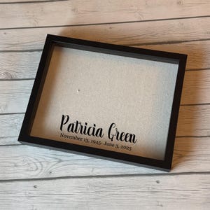 Puede incluir: Un marco negro con el nombre "Patricia Green" y las fechas "November 13, 1945- June 3, 2025" impresos en negro. El marco tiene un respaldo de tela de color claro.