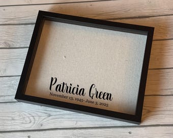 Peut inclure: Un cadre noir avec le nom "Patricia Green" et les dates "November 13, 1945- June 3, 2025" imprimés en noir. Le cadre est doté d'un fond en tissu de couleur claire.