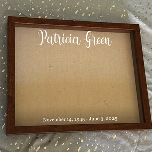 Puede incluir: Una caja de sombra rectangular con marco de madera oscura y respaldo de tela beige. El nombre "Patricia Green" está escrito en blanco en la parte superior. Las fechas "November 14, 1945 - June 3, 2025" están impresas en blanco en la parte inferior.