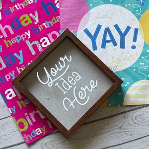 Puede incluir: Una caja de sombra cuadrada de madera con el texto "Your Idea Here" en escritura blanca. La caja se coloca sobre un fondo de papel de regalo de cumpleaños y una bolsa de regalo con la palabra "YAY!" en azul.