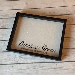 Puede incluir: Un marco negro con el nombre "Patricia Green" y las fechas "13 de noviembre de 1945 - 3 de junio de 2025" impresos en negro sobre un fondo claro. El marco está inclinado sobre una superficie de madera clara.