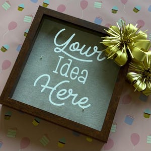 Puede incluir: Un marco de sombra de madera marrón oscuro con el texto "Your Idea Here" en escritura blanca. El fondo es de tela gris claro. Lazos de cinta dorada en la esquina superior derecha. El fondo es rosa con ilustraciones de pasteles y globos.