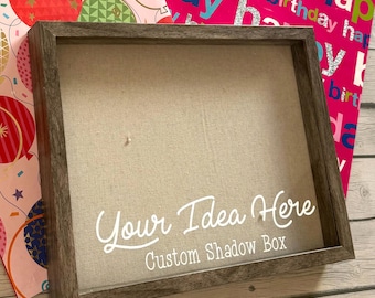 Peut inclure: Une boîte à ombre en bois avec un cadre brun clair et un fond en tissu beige. La boîte a un devant en verre transparent et le texte "Your Idea Here Custom Shadow Box" est imprimé sur le devant.