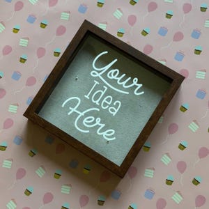 May include: A dark brown shadow box with the words "Your Idea Here" in white script. The box has a clear front and is set against a pink background with cupcake and balloon patterns.
