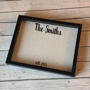May include: A black framed shadow box with a light-colored fabric backing. The words "The Smiths" are written in a cursive font at the top, with "est. 2025" at the bottom. The frame is rectangular.