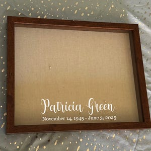 Puede incluir: Una pieza conmemorativa enmarcada con un marco de madera marrón. El fondo es de tela beige. El nombre "Patricia Green" está escrito en blanco, junto con las fechas "14 de noviembre de 1945 - 3 de junio de 2025".