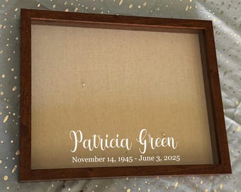 Peut inclure: Un cadre commémoratif avec un cadre en bois marron. Le fond est en tissu beige. Le nom "Patricia Green" est écrit en blanc, avec les dates "14 novembre 1945 - 3 juin 2025".