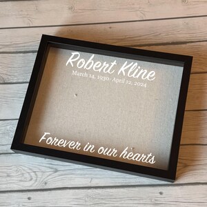Puede incluir: Una caja de sombra con marco negro con el texto "Robert Kline" y las fechas 14 de marzo de 1930 - 12 de abril de 2024, en escritura blanca. La frase "Forever in our hearts" también está en escritura blanca. El fondo es de tela beige claro.