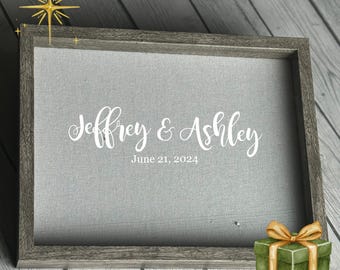 May include: A framed gray canvas with the names "Jeffrey & Ashley" and the date "June 21, 2024" in white script. A small green gift box with a gold bow sits in the corner. The frame is gray.