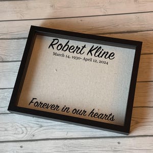 Puede incluir: Un marco negro con el nombre "Robert Kline" y las fechas "14 de marzo de 1930 - 12 de abril de 2024" en escritura negra. La frase "Forever in our hearts" también está en escritura negra.