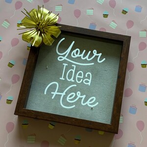Puede incluir: Un marco de sombra cuadrado de madera marrón oscuro con las palabras "Your Idea Here" en escritura blanca. Un lazo dorado está unido a la esquina superior izquierda. El fondo es rosa con patrones de pastel y globos.