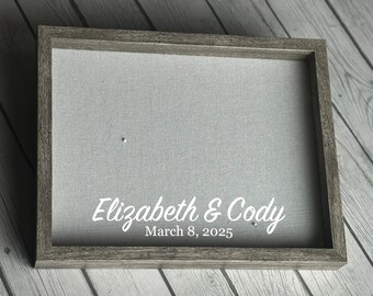 May include: A gray framed sign with a light gray fabric background. The sign has the names "Elizabeth & Cody" and the date "March 8, 2025" in white script. The frame is a weathered gray color.