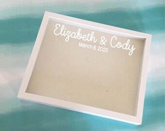 May include: A white framed shadow box with the names "Elizabeth & Cody" and the date "March 8, 2025" in white script. The interior of the box has a beige background.