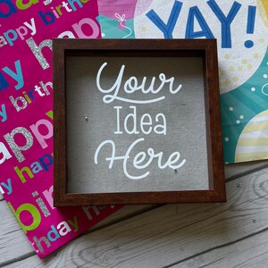 Puede incluir: Un letrero cuadrado con un marco de madera oscura y el texto blanco "Your Idea Here". El letrero tiene un respaldo de tela neutra. Se muestra sobre una superficie de madera con bolsas de regalo coloridas.