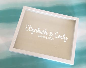 May include: A white framed sign with the names "Elizabeth & Cody" and the date "March 8, 2025" in white script. The background is a light tan color, and the frame is rectangular.