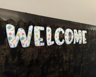 Może przedstawiać: Czarna tablica z napisem "WELCOME" napisanym dużymi, kolorowymi literami. Litery są ozdobione wzorem w szkolnym stylu z jabłkami, ołówkami i żarówkami.