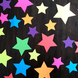 May include: A collection of colorful paper stars in various shades of blue, green, orange, pink, purple, and yellow. The stars are arranged on a dark brown wooden surface.