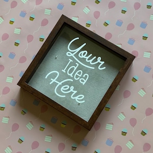 Puede incluir: Un marco de sombra marrón oscuro con las palabras "Your Idea Here" en escritura blanca. La caja tiene una parte delantera transparente y está colocada sobre un fondo rosa con patrones de cupcakes y globos.