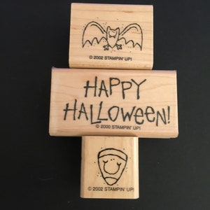 May include: Three wooden stamps with Halloween themes. The top stamp features a black bat with white eyes. The middle stamp says "HAPPY HALLOWEEN!" and the bottom stamp features a smiling orange pumpkin.