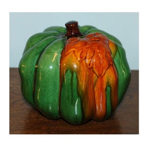 May include: A ceramic pumpkin with a glossy finish. The pumpkin is primarily green with a section of orange and brown glaze. The stem is a dark brown color. The pumpkin is sitting on a wooden surface.