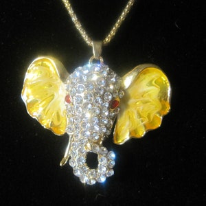 May include: A gold chain necklace with a yellow enamel elephant pendant. The elephant is decorated with clear rhinestones and has red eyes.