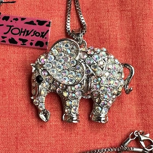 May include: A silver-toned elephant pendant necklace featuring iridescent rhinestones covering the body and a large heart-shaped ear. The elephant has a single black eye and is attached to a box chain. A pink tag with black animal print and the text JOHNSON is visible.