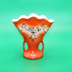 May include: Orange ceramic vase with a scalloped rim and floral design. The vase has two handles and a white background with green and orange flowers.