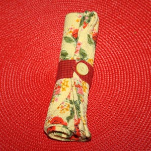 May include: Rolled floral napkin with a red and white checkered band secured by a large, round, beige button. The napkin features a pattern of red, pink, yellow, and green flowers on a cream background. The napkin is on a red woven placemat.