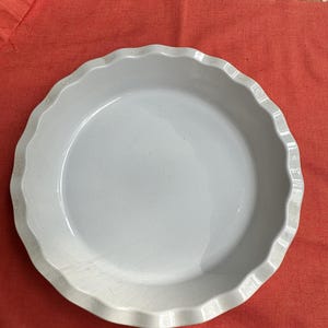 May include: A round white ceramic pie dish featuring a smooth interior surface and a decorative scalloped edge. The dish has a glossy finish and a consistent circular shape, designed for baking purposes with a wide, open basin and fluted rim.