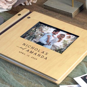 May include: A light wood wedding photo album with a framed photo of a couple. The album is open, displaying the names "Nicholas and Amanda" and the wedding date "April 13, 2025". A Fujifilm Instax camera and several instant photos are nearby.