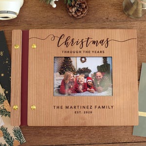 May include: A wooden photo album with the words "Christmas Through The Years" and a family photo. The album is surrounded by Christmas cards, a gift card, and festive decorations. The family name is "The Martinez Family" and the year is "EST. 2020".