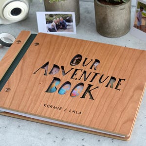 May include: A wooden photo album with the words "OUR ADVENTURE BOOK" cut out of the cover. The album has a green spine and is open to reveal photos. The names "KERMIE & LALA" are printed below the title. A white instant camera and other travel-themed items are in the background.