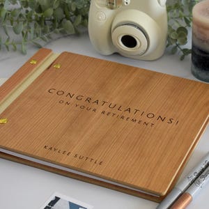 以下が含まれることがあります： 「CONGRATULATIONS! ON YOUR RETIREMENT」の文字が刻まれた木製のフォトアルバム。アルバムは明るい茶色で、金色の金具が付いています。メタリックなSharpieマーカー2本と、富士フイルムのインスタントカメラも写っています。