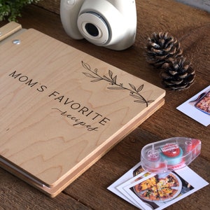 May include: A wooden recipe book with the text "MOM'S FAVORITE recipes" on the cover. The book is closed and lying on a wooden surface. There is a white instant camera, two pine cones, and a roll of tape on the surface.