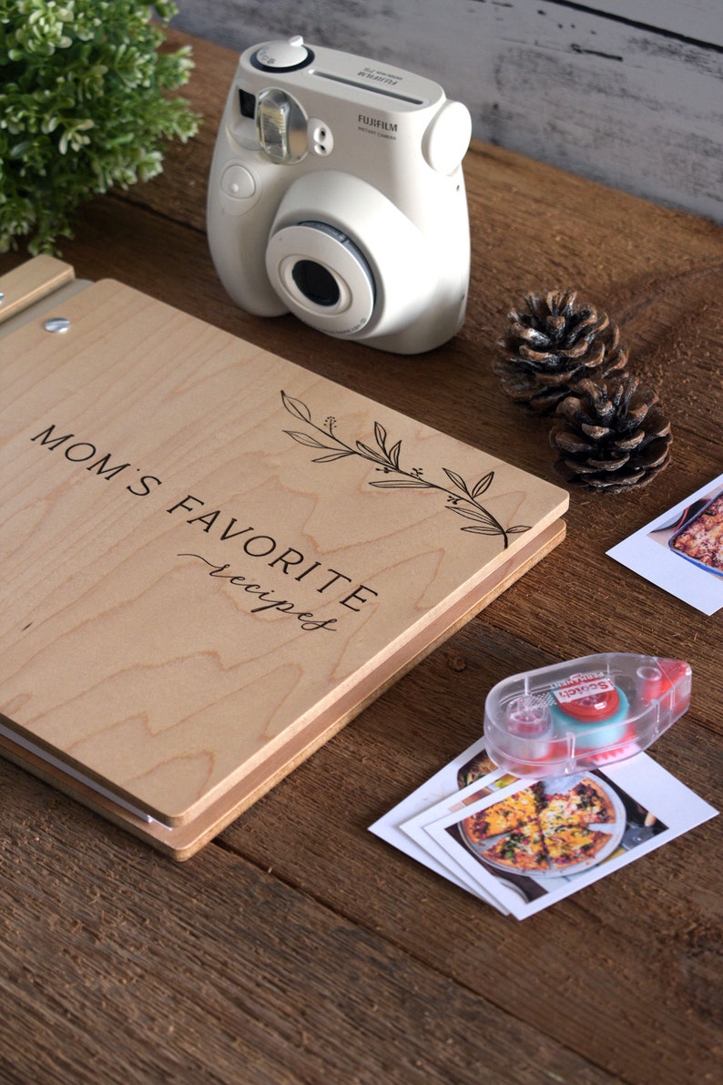 May include: A wooden recipe book with the text "MOM'S FAVORITE recipes" on the cover. The book is closed and lying on a wooden surface. There is a white instant camera, two pine cones, and a roll of tape on the surface.
