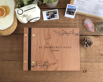 May include: A rectangular wooden guestbook with a light brown wood grain finish. The cover features engraved black text reading 88 TIMBERCREST ROAD Guestbook, accented by two delicate black line-art botanical branch illustrations at the top and bottom.