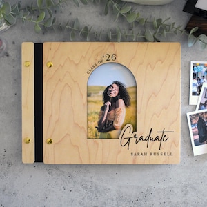 a wooden graduation scrapbook with a photo of a smiling young woman, surrounded by various items such as a pen, a small picture frame, and some plants.