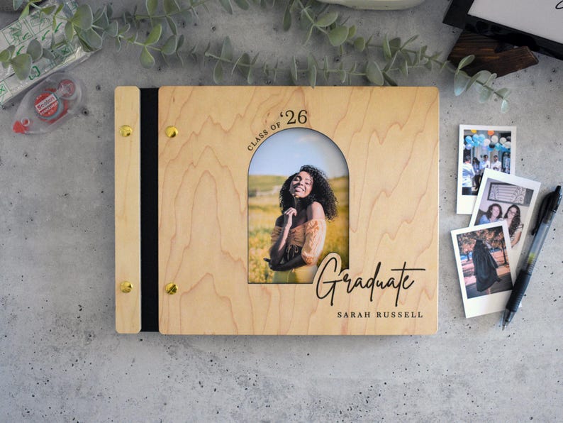 a wooden graduation scrapbook with a photo of a smiling young woman, surrounded by various items such as a pen, a small picture frame, and some plants.