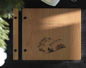 May include: A rectangular wooden guest book with a light brown finish, featuring a black engraved mountain landscape and cabin illustration. The text reads: welcome to ELK RIDGE CABIN in Pray, Montana. It has a side binding strip with two black screws.