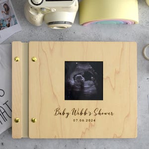 May include: A light-colored wooden photo album with gold-toned metal binding screws. The cover features a square cutout displaying a black and white ultrasound image, with the printed text Baby Webb's Shower and the date 07.06.2024 below it.