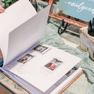 May include: A white guest book with a brown cover is open to reveal pages with photos and handwritten messages. The book is on a table with a light blue tablecloth and a copper framed sign that says "newlywed" in white cursive.