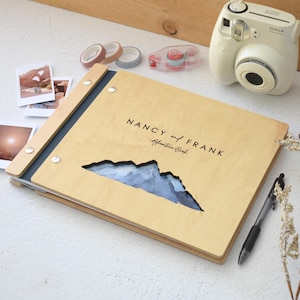 May include: A light-coloured wooden photo album with a dark blue spine and silver metal fasteners. The cover features a mountain-shaped cutout revealing a blue landscape and the text NANCY and FRANK Adventure Book printed in black ink.