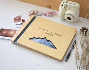 May include: A light-colored wooden photo album with a dark blue spine and silver metal fasteners. The cover features a mountain-shaped cutout revealing a blue landscape and the text NANCY and FRANK Adventure Book printed in black ink.
