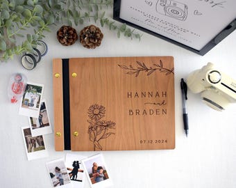 May include: A rectangular wooden photo album with a light brown finish, featuring a laser-engraved design of two daisies and a leafy branch. The text reads "HANNAH and BRADEN" and "07.12.2024". The album is bound with gold-coloured metal screws.
