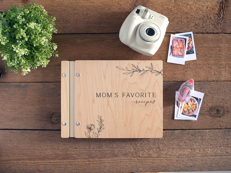 May include: A wooden recipe book with a light wood finish and a black inscription that reads "Mom's Favorite Recipes". The book has a decorative floral design and a metal ring binder.