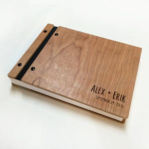 May include: A rectangular wooden photo album with a light brown wood grain finish. The cover features the engraved text ALEX + ERIK and SEPTEMBER 21, 2016. The album is bound with a black spine and secured by two black metal rivets on the left side.