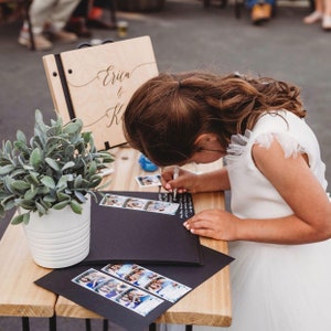 May include: A young girl in a white dress writes in a black photo album with white pages. The album is on a wooden table with a potted plant and a wooden sign that says "Erica & K" in cursive.