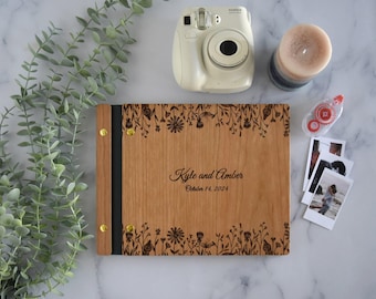 Könnte beinhalten: Ein rechteckiges Fotoalbum aus Holz mit schwarzem Buchrücken und goldfarbenen Metallverschlüssen. Der Einband zeigt laser-gravierte Blumenränder und den Text "Kyle and Amber" über dem Datum "October 14, 2024" mittig auf der Holzfront.