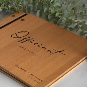May include: A rectangular wooden book with a light brown grain, featuring a dark brown binding strip secured by two metal rivets. The cover is engraved with the word Officiant, the name Lanie Dornie, and the text Michael + Krystan 12.09.2023.